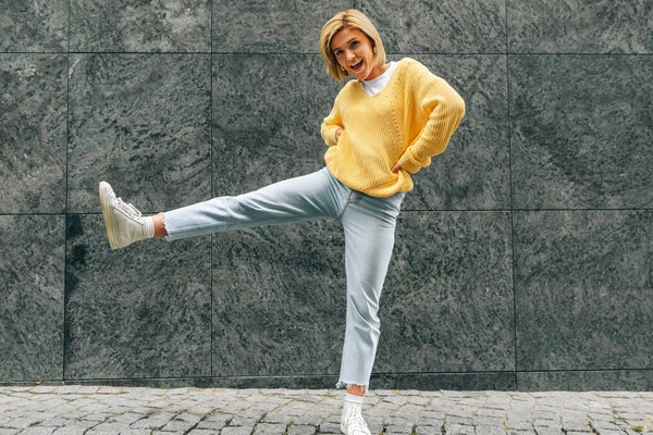 Horizontal outdoor image of attractive young woman wearing yellow sweater, blue hight waist jeans, jumping high, feeling happy on the city street. Student blonde female as joyful expression