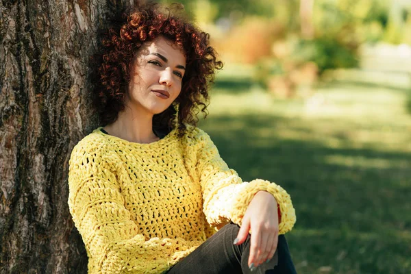 Εξωτερική οριζόντια εικόνα της όμορφης νεαρής γυναίκας με σγουρά μαλλιά και χαμόγελο. Θηλυκό έχει θετική έκφραση, φοράει πλεκτό κίτρινο πουλόβερ και ποζάρει στο φόντο της φύσης. Άνθρωποι, τρόπος ζωής — Φωτογραφία Αρχείου