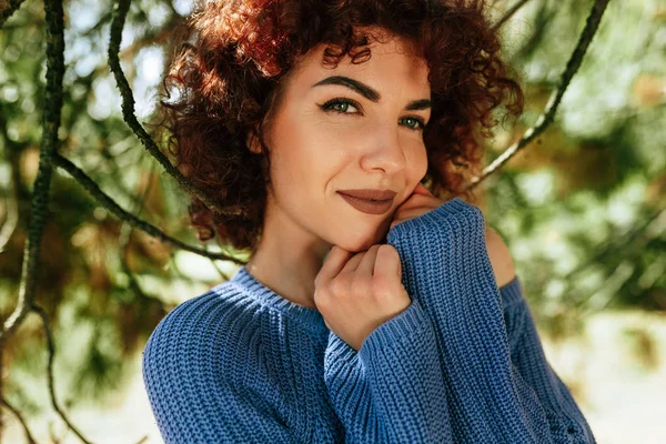 Outdoor close-up portrait of gorgeous young woman with curly hair, smiling and looking to the camera, posing against nature background, has positive expression, wearing blue knitted sweater. — Stock Photo, Image