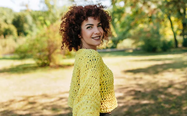 Красива молода жінка з кучерявим волоссям, посміхаючись, має позитивний вираз, одягнена в в'язаний жовтий светр і позує на фоні природи. Люди і спосіб життя — стокове фото