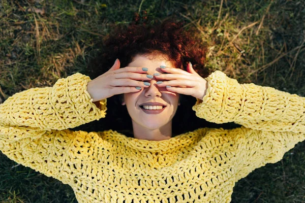 Вид зверху красива молода жінка з кучерявим волоссям, посміхаючись і покриваючи очі руками, лежить на траві, має позитивний вираз, одягнений в в'язаний жовтий светр. Люди і спосіб життя — стокове фото