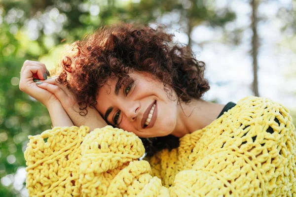Κοντινό πλάνο εξωτερικό πορτρέτο της ελκυστικής νεαρής γυναίκας με σγουρά μαλλιά και χαμόγελο. Θηλυκό έχει θετική έκφραση, φοράει πλεκτό κίτρινο πουλόβερ και ποζάρει στο φόντο της φύσης. Άνθρωποι, τρόπος ζωής — Φωτογραφία Αρχείου
