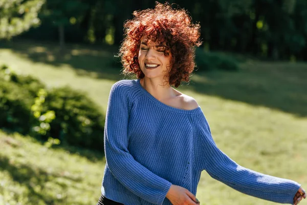 Retrato horizontal ao ar livre de bela jovem com cabelo encaracolado, sorrindo e apreciando o tempo, posando contra o fundo da natureza, tem expressão positiva, vestindo camisola de malha azul . — Fotografia de Stock