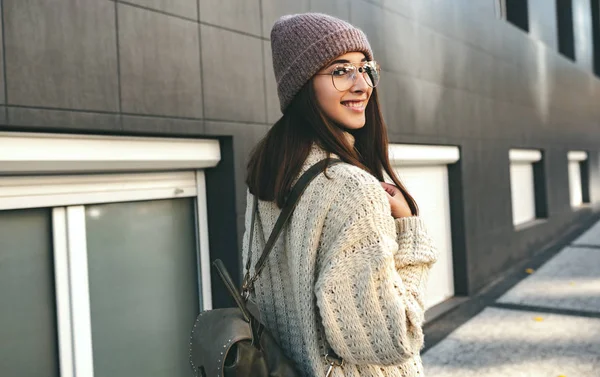 Vista trasera de la imagen de una bonita mujer sonriente mirando hacia atrás, caminando por la calle, usando suéter de punto y gafas graduadas. Hermosa joven con mochila esperando a sus amigos en la avenida . —  Fotos de Stock