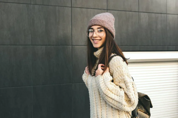 Retrato horizontal de una joven feliz sonriente mirando a la cámara, caminando por la calle de la ciudad, vistiendo suéter de punto, anteojos transparentes y sombrero . —  Fotos de Stock