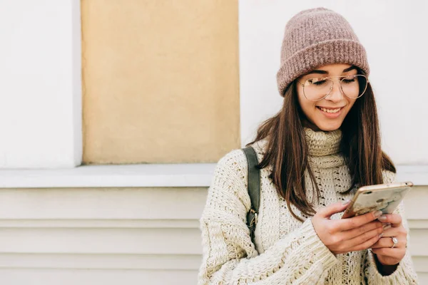 Attractive young woman standing outdoors against building's wall, playing on smart phone. Brunette female dressed in knitted sweater, hat, eyeglasses send messages from her device on the city street — ストック写真
