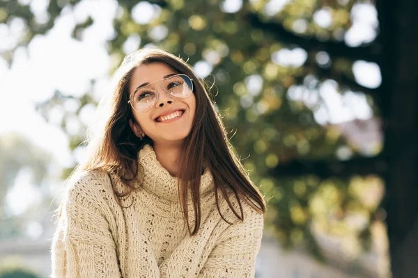 Szczęśliwa studentka uśmiechnięta, ubrana w sweter z dzianiny i modne przezroczyste okulary. Piękny szczęśliwy młoda kobieta pozowanie na zewnątrz na słońcu na ulicy miasta. — Zdjęcie stockowe