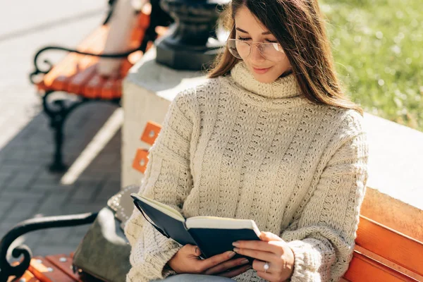 Fotografia aérea de uma jovem mulher usando suéter e óculos transparentes, sentada ao ar livre no banco e lendo o livro enquanto espera seus amigos. Jovem estudante aprendendo na rua da cidade . — Fotografia de Stock