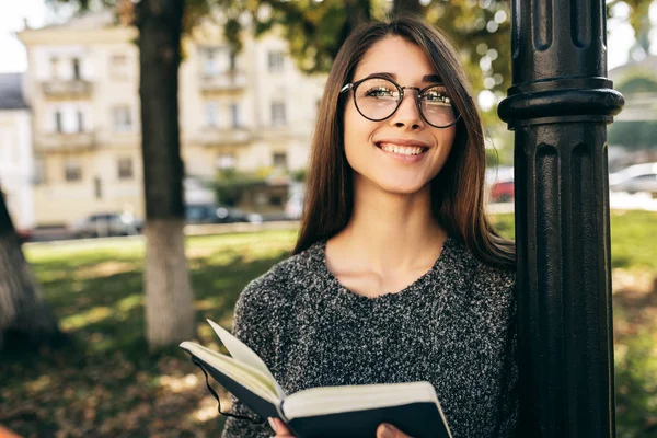 Portrét krásné mladé ženy ve svetru a průhledných brýlích, stojící venku na městské ulici a čtoucí knihu, zatímco čeká na svého přítele. Ženské studentské vzdělávání ve městě ulici — Stock fotografie