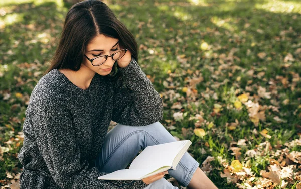Atraktivní mladá žena ve svetru a průhledných brýlích, stojí venku na městské ulici a čte si knihu, zatímco čeká na svého přítele. Ženské studentské vzdělávání ve městě ulici — Stock fotografie