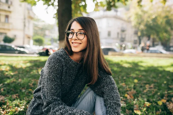 Imagem ao ar livre de bela jovem sorridente sentada na grama verde no parque da cidade, vestindo suéter de malha e óculos transparentes. bela jovem mulher descansando fora — Fotografia de Stock