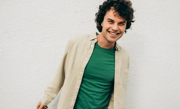 Jovem bonito posando para propaganda, sorrindo e olhando para a câmera, de pé na construção de parede de concreto ao ar livre. Retrato de estudante inteligente feliz macho com cabelo encaracolado — Fotografia de Stock