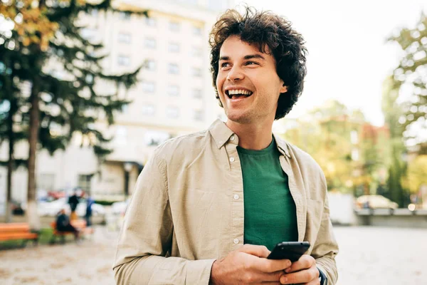Счастливый улыбающийся молодой человек, стоящий на улице, имеет радостное выражение во время игры на мобильном телефоне. Молодой красивый мужчина с кудрявыми волосами просматривает онлайн на своем смартфоне на городской улице . — стоковое фото