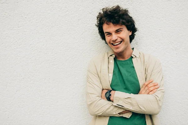 Bonito jovem posando para propaganda, sorrindo e olhando para a câmera, de pé na construção de parede de concreto ao ar livre. Retrato de estudante inteligente feliz macho com cabelo encaracolado com braços cruzados . — Fotografia de Stock