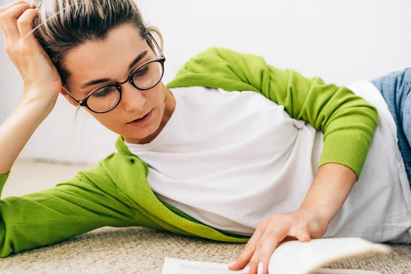 Imagem de close-up de jovem inteligente lendo livro, vestindo casaco verde e t-shirt branca, óculos, deitado no tapete. Feminino relaxante durante a leitura de um livro. Estudante menina estudando em casa . — Fotografia de Stock