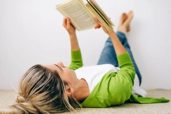 Jovem mulher lendo livro, vestindo óculos transparentes deitado no tapete na sala. Feminino relaxante na acolhedora sala de estar, lendo um livro. Estudante menina sentado em casa e estudando . — Fotografia de Stock