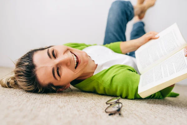 Kitap okuyan, yeşil hırka ve beyaz tişört giyen, odada halıya uzanmış mutlu genç bir kadın. Kadın dinleniyor ve kitap okuyor. Öğrenci kızın evde çalışmasının neşeli bir ifadesi var. — Stok fotoğraf