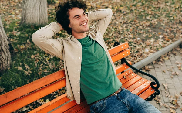 Au-dessus de la vue de beau jeune homme aux cheveux bouclés, souriant et levant les yeux, se sentant joyeux, assis sur le banc dans la rue de la ville à l'extérieur. heureux intelligent étudiant mâle a rêver expression . — Photo