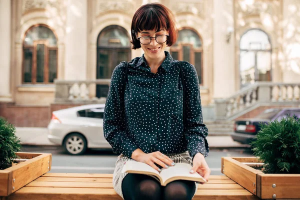 Горизонтальне зображення розумної молодої жінки в зеленій сорочці з білими крапками і прозорими окулярами, сидячи на лавці на вулиці міста і читаючи книгу. Молода студентка вчиться на вулиці — стокове фото