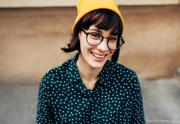 Ao ar livre close-up retrato de bela jovem mulher sorrindo amplamente vestindo camisa verde com pontos brancos, óculos transparentes e chapéu amarelo. Jovem estudante posando na rua da cidade . — Fotografia de Stock
