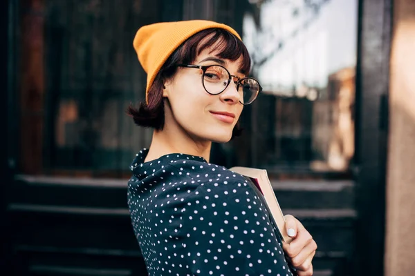 Портрет розумної молодої студентки в жовтому капелюсі, прозорих окулярах і зеленій сорочці з білими крапками з книгами в руках, що стоять проти університету . — стокове фото