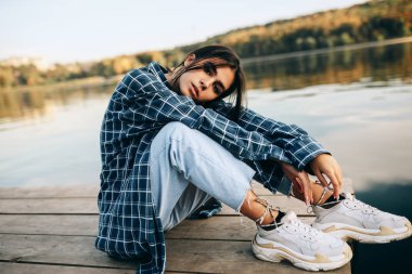 Güzel esmer bir kadının yatay görüntüsü, günlük moda kıyafetler giymiş, doğa arka planında oturuyor. Gölde poz veren güzel bir kadın, rüya gibi bir ifadesi var..