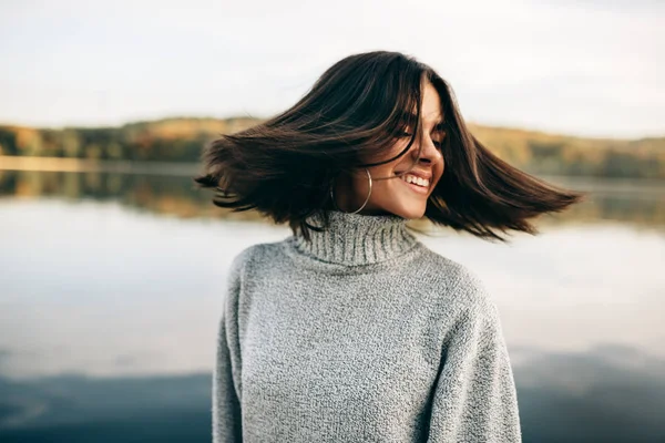 Close-up ao ar livre retrato de feliz jovem morena com olhos fechados com cabelo soprando, vestindo camisola cinza, posando no fundo da natureza. Bonita fêmea posando contra o lago . — Fotografia de Stock