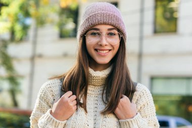 Gülümseyen güzel kız öğrencinin portresi doğrudan kameraya bakıyor, şehrin caddesinde duruyor, bahar zamanı örülü süveter giyiyor. Sırt çantalı, dışarıda poz veren güzel bir kadın.