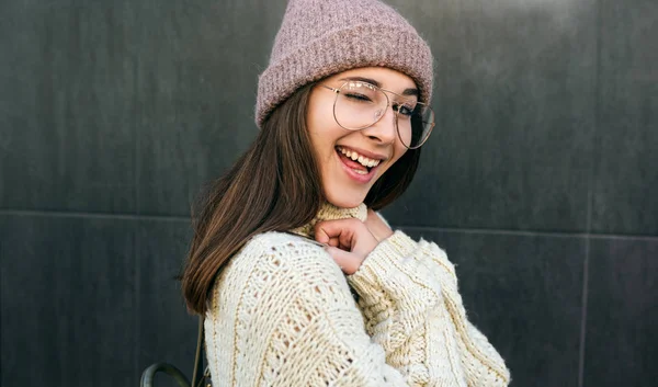 Funny smiling young woman blink with an eye and looking at the camera, wearing knitted sweater, transparent eyeglasses and hat, posing on the city street. — 스톡 사진