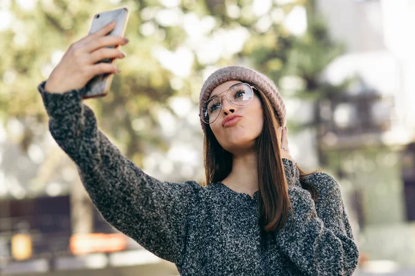 Jovem mulher de pé ao ar livre e tomando auto retrato no telefone inteligente. Morena mulher vestida com camisola de malha, chapéu, óculos fazendo selfie em seu dispositivo na rua da cidade . — Fotografia de Stock