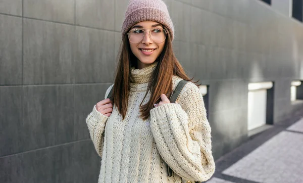 Mujer joven sonriente caminando por la calle de la ciudad, vistiendo suéter, sombrero rosa y gafas transparentes. Estudiante bastante femenina que va a la Universidad . —  Fotos de Stock