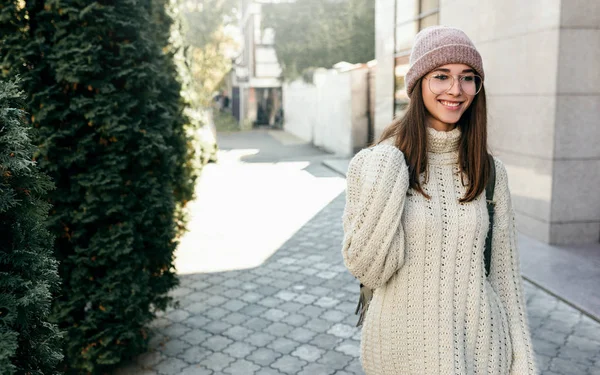 Imagem da bela jovem mulher sorrindo amplamente e olhando diretamente para a câmera, andando na rua da cidade, vestindo suéter, chapéu rosa e óculos transparentes da moda . — Fotografia de Stock