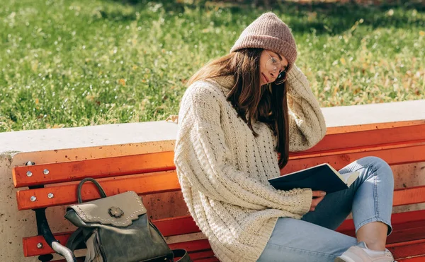Mladá žena ve svetru, klobouku a průhledných brýlích seděla na lavičce na městské ulici a četla knihu, zatímco čekala na své přátele. Mladá studentka se učí venku. — Stock fotografie