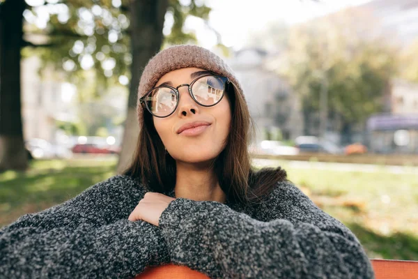 Portrét krásné mladé ženy, usmívající se, dívající se do kamery, s módními průhlednými brýlemi, svetrem a kloboukem. Atraktivní žena sedí a odpočívá na lavičce v parku. — Stock fotografie