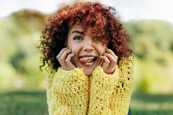 Retrato de cerca de una joven divertida con el pelo rizado mostrando la lengua y sonriendo. La hembra tiene expresión positiva, vistiendo suéter amarillo y posando sobre el fondo de la naturaleza. Personas, estilo de vida —  Fotos de Stock