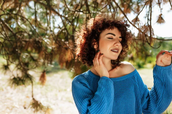 Beautiful young woman with curly hair, smiling and enjoying the weather, posing against nature background, has positive expression. — Stock Photo, Image