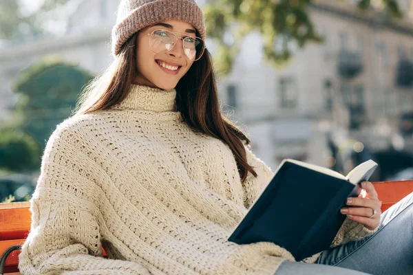 Sorrindo jovem inteligente vestindo suéter, chapéu e óculos transparentes, sentada no banco na rua da cidade, lendo o livro enquanto espera seus amigos. Estudante muito feminino aprendendo fora — Fotografia de Stock