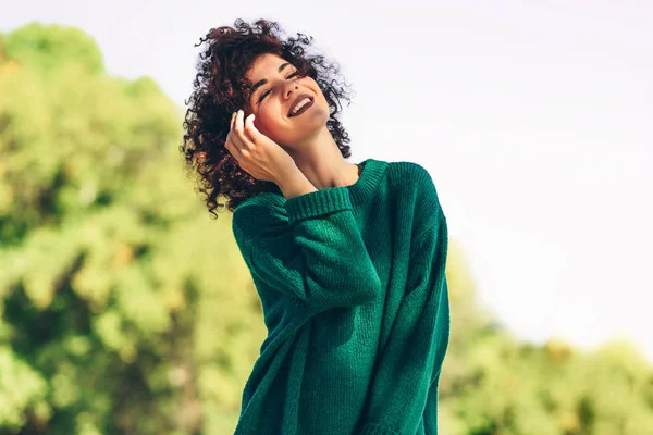 Beeld van gelukkige mooie jonge vrouw lachend poserend tegen de achtergrond van de natuur met winderig krullend haar, hebben positieve uitdrukking, dragen in groene trui. Mensen, reizen en levensstijl. — Stockfoto