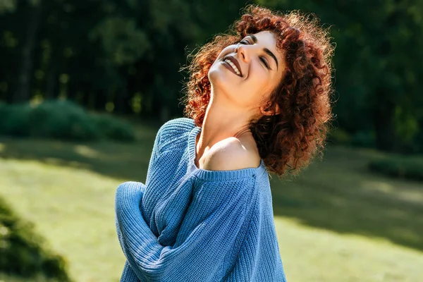 Красива молода жінка з кучерявим волоссям, посміхаючись і насолоджуючись погодою, позує на фоні природи, має позитивний вираз, одягнений в синій светр . — стокове фото