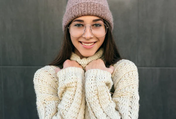 Retrato de bela jovem sorridente posando na rua da cidade contra a parede cinza, usando suéter, chapéu rosa e óculos transparentes. Estudante muito feminina indo para a faculdade . — Fotografia de Stock