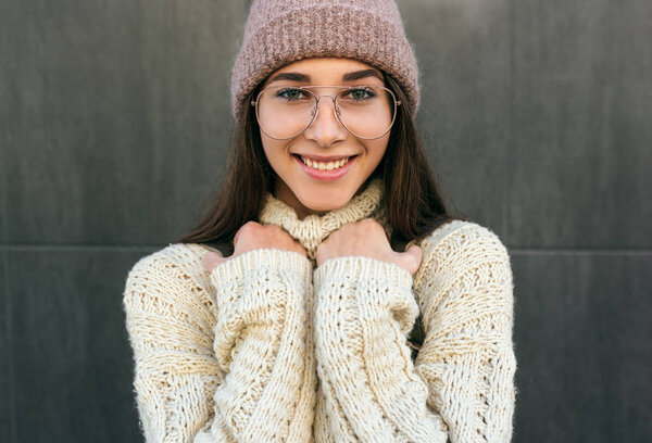 Portrait of beautiful smiling young woman posing in the city street against grey wall, wearing sweater, pink hat and transparent eyeglasses. Pretty female student going to the college. — Stock Photo, Image