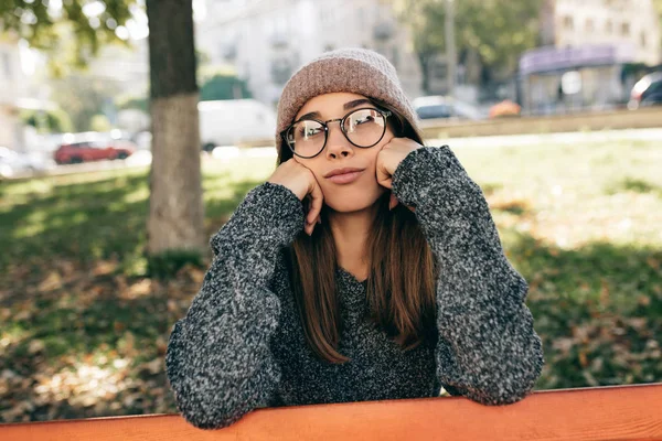 Smutna młoda kobieta patrząca w kamerę, nosząca modne przezroczyste okulary, sweter i kapelusz. Piękna kobieta siedzi i relaksuje się na ławce w parku. — Zdjęcie stockowe