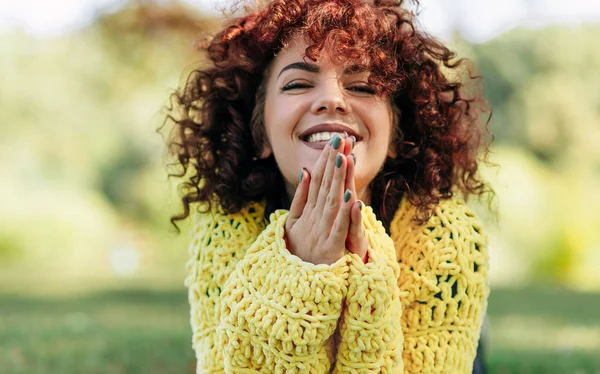 Portrait of cheerful young woman with curly hair smiling broadly with toothy smile. Female has positive expression, wearing yellow sweater and posing against nature background. People, lifestyle — 스톡 사진