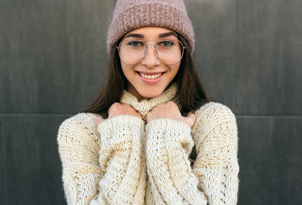 Portrait of beautiful smiling young woman posing in the city street against grey wall, wearing sweater, pink hat and transparent eyeglasses. Pretty female student going to the college. Stock Image