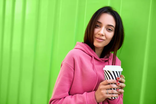 Élégante jeune femme heureuse portant un sweat rose tenant sur la route. Portrait de fille souriante buvant du café à l'extérieur sur le fond vert — Photo