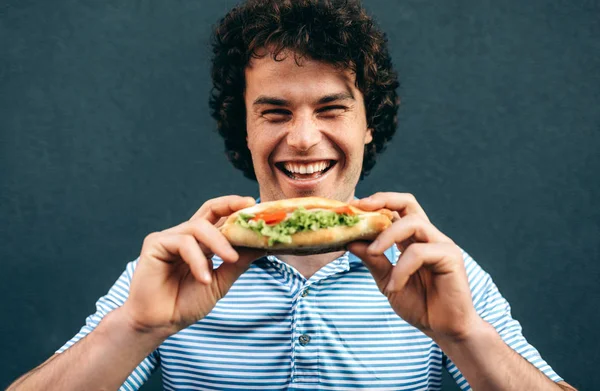 Egy közeli kép fiatal boldog emberről, aki egészséges hamburgert eszik. Jóképű férfi egy gyorsétteremben hamburgert eszik a szabadban. Férfi göndör hajjal, utcai kajával és hamburgerrel. — Stock Fotó