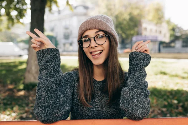 Śmieszna młoda kobieta uśmiechnięta, nosząca sweter i modne przezroczyste okulary pokazujące gest spokoju. Piękna szczęśliwa młoda kobieta relaksująca się na zewnątrz na słońcu w parku miejskim. — Zdjęcie stockowe