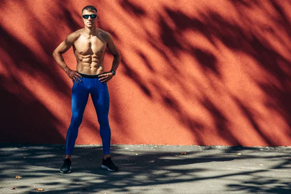 Homem atleta jovem posando ao ar livre contra a parede de concreto vermelho durante o treino. Atleta masculino em óculos escuros pretos descansando após o treino fora . — Fotografia de Stock