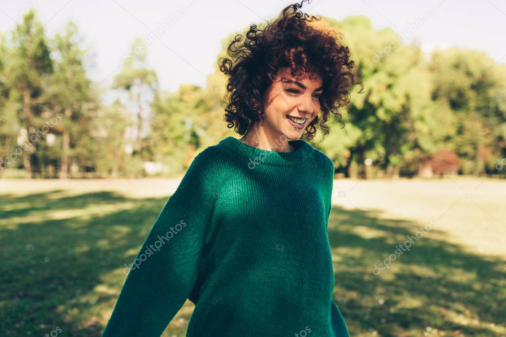 Beautiful young woman smiling posing against nature background with windy curly hair, have positive expression, wearing in green sweater. People, travel and lifestyle.