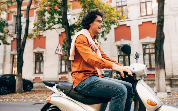 Imagem de jovem inteligente dirigindo em sua scooter, vestido com roupa casual e óculos transparentes, com cabelo encaracolado, tem expressão alegre . — Fotografia de Stock